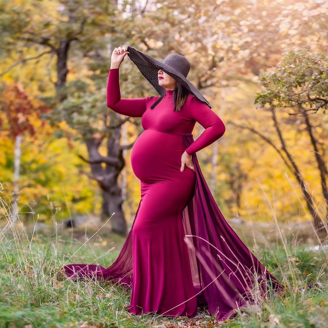 En gravid kvinna i röd klänning med långt släp står i på en äng med höstfärgade träd runt. På sig har hon en stor svart hatt.