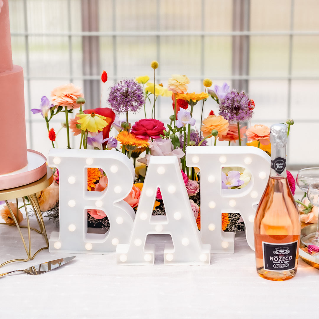 Stora bokstäver med lampor står på ett dukat bord och stavar ut ordet BAR. På bordet syns också en bröllopstårta, bubbel i flaska och ett färgglatt blomsterarrangemang.