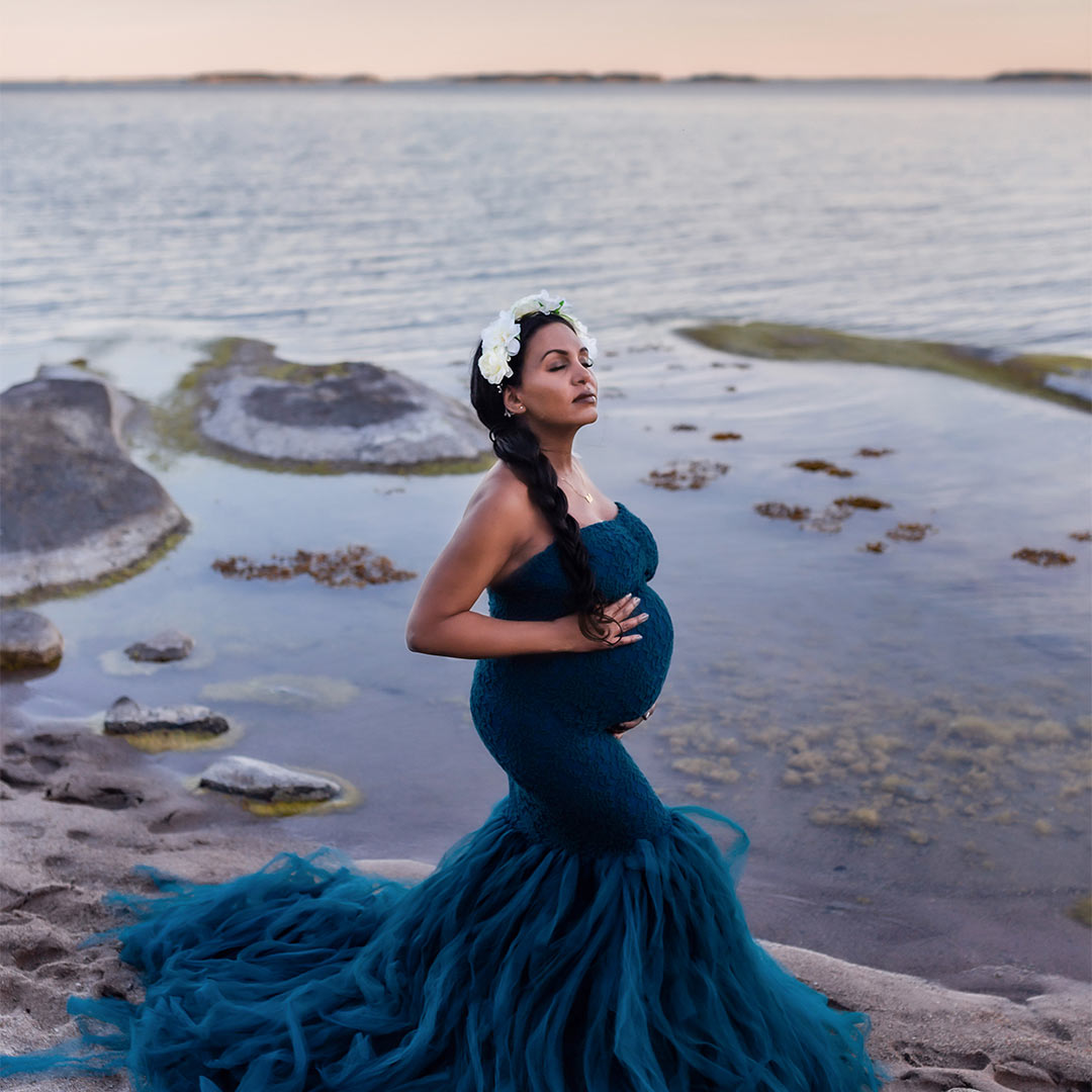 Gravidfoto i blå toner med havet i bakgrunden. Den vackra gravida kvinnan har en blå tyllklänning och vit blomkrans.