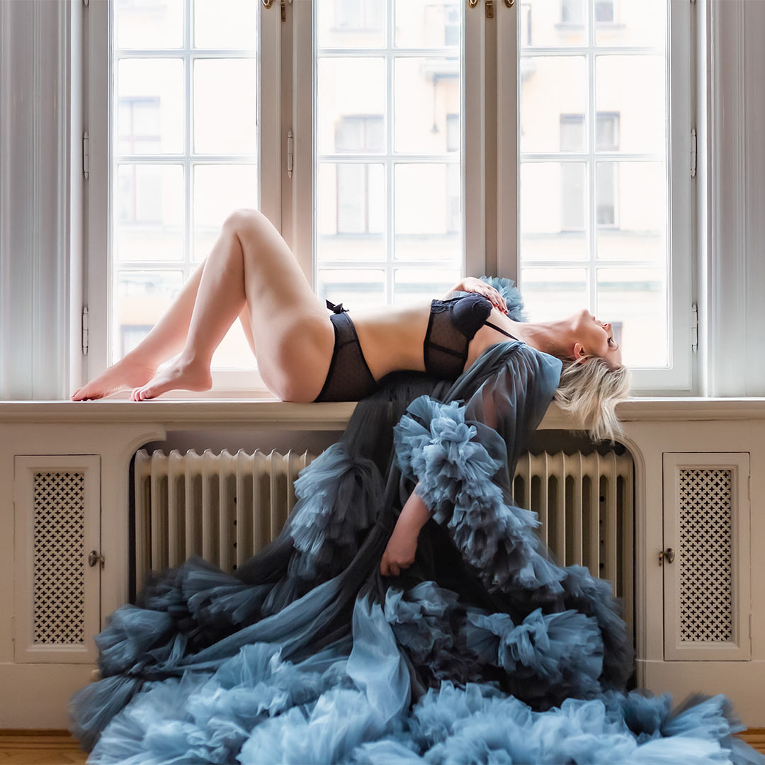 Boudoirfotografering av en kvinna som ligger i ett fönster och har på sig svarta underkläder och en blå tyllklänning.
