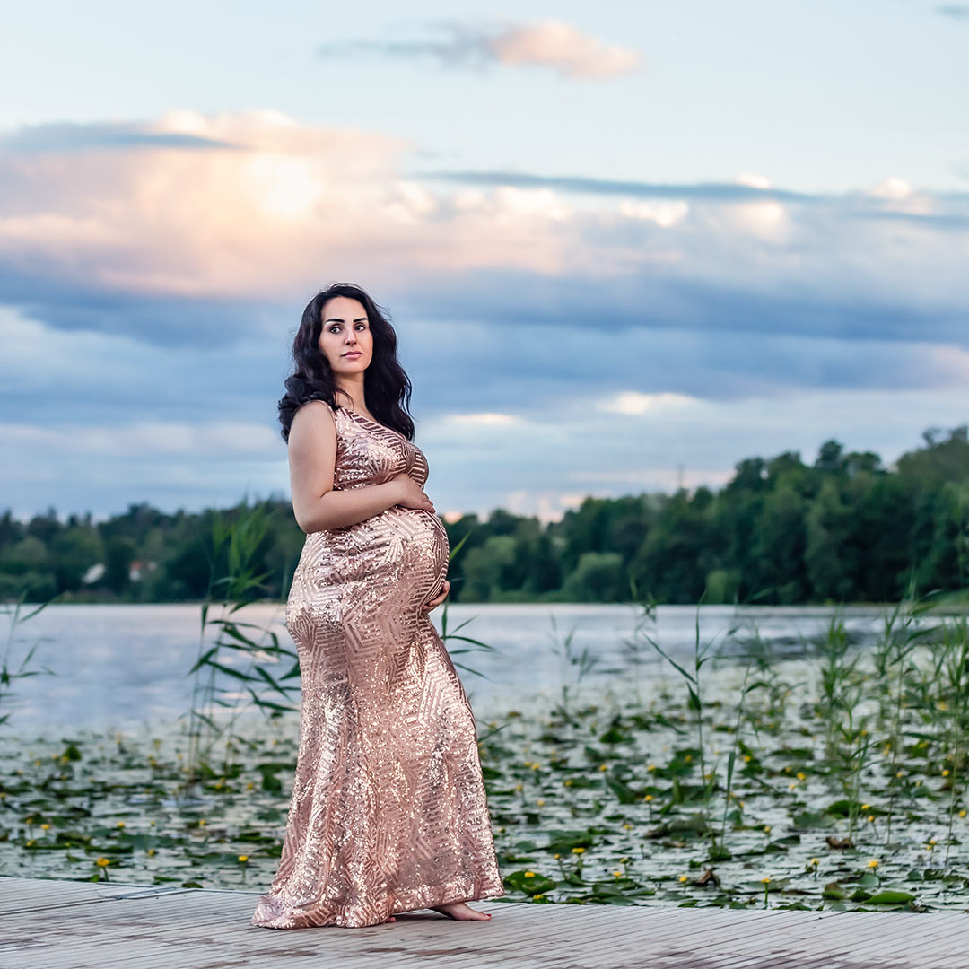 En gravid kvinna i rosaglittrande gravidklänning går på en brygga. Himlen är storslaget vacker med stora moln och på vattnet växer näckrosor.