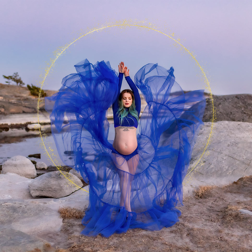 En gravid kvinna dansar så att den blå tyllkjolen flyger, på klipporna vid havet.