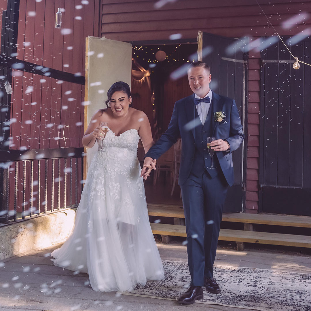 Ett nyvigt bröllopspar går ut från ett rödmålat hus medan deras nära och kära kastar ris. De skrattar och håller i champagneglas.