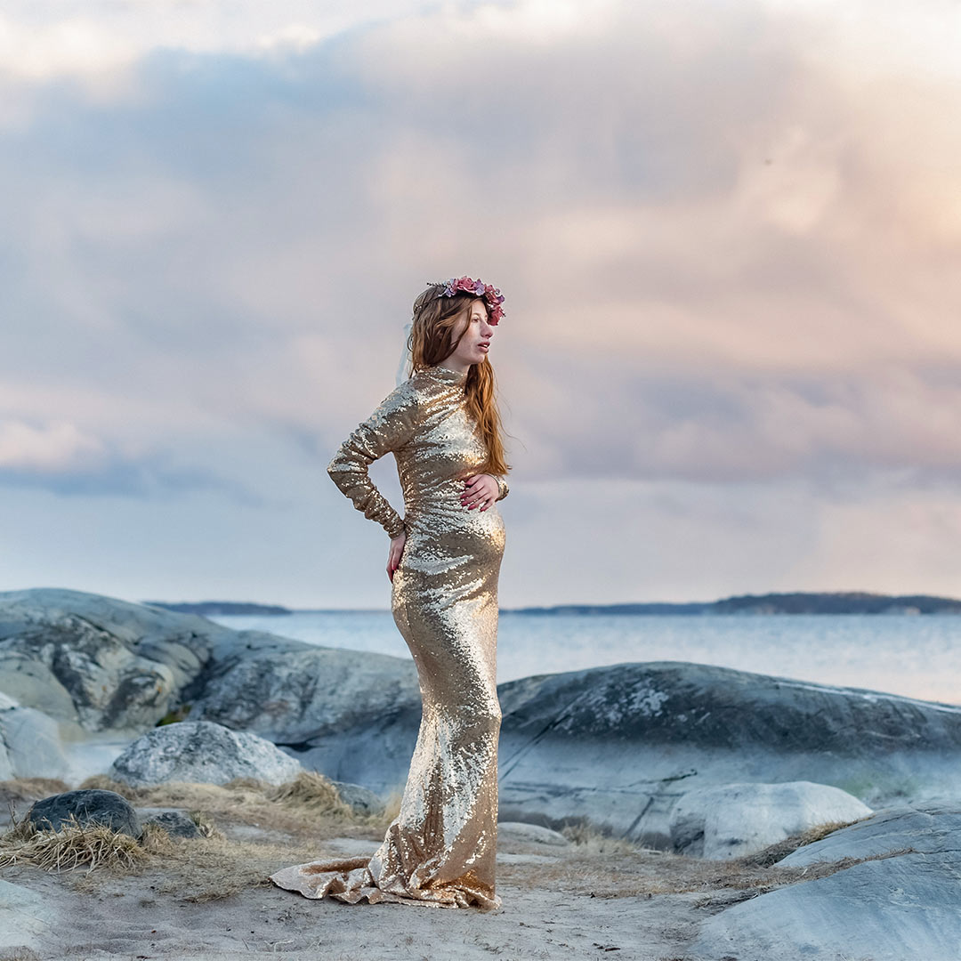 Gravidfotografering vid havet där den gravida kvinnan står bland klipporna iförd en guldglittrande klänningen. Himlen har storslagna moln färgade av solnedgången.