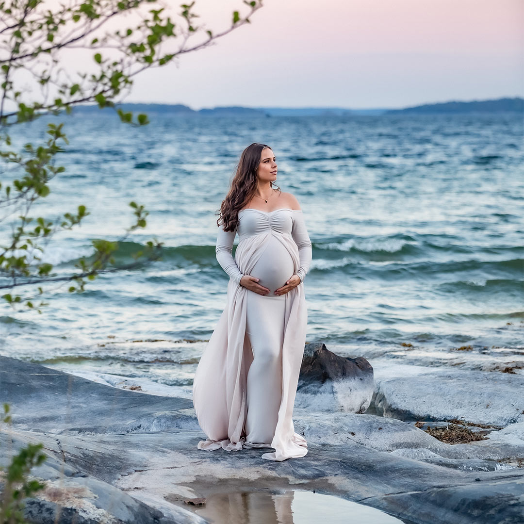 Gravidfotografering vid havet och klipporna i kvällsljus. Kvinnan har en beige gravidklänning med släp.