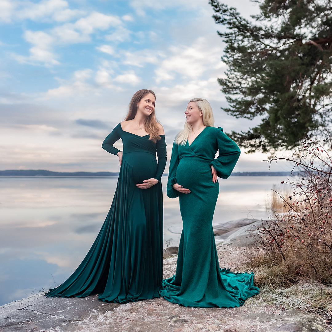 Gravidfotografering av två gravida systrar som står på klippa framför ett stilla hav. De tittar på varandra och ler.