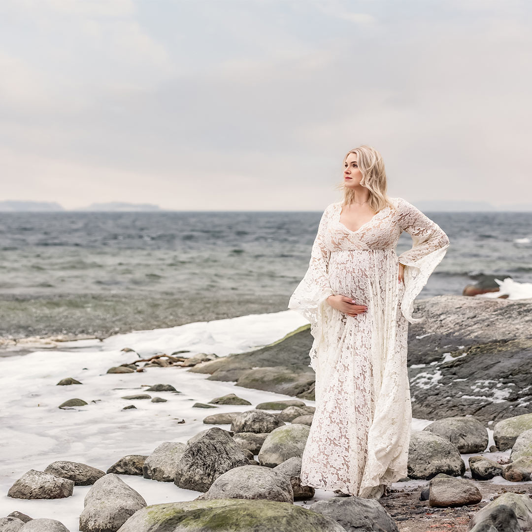 En gravid kvinna i vit spetsklänning står vid ett isande kallt hav.