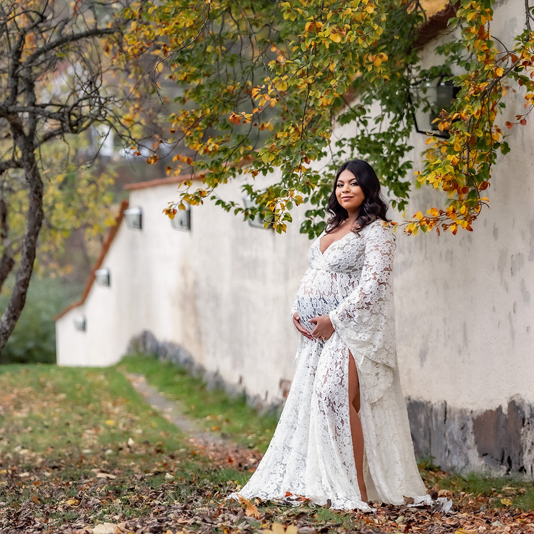 En gravid kvinna står vid en vit mur i en vit spetsklänning. Det är höst och grenarna är fulla av höstfärgade löv.