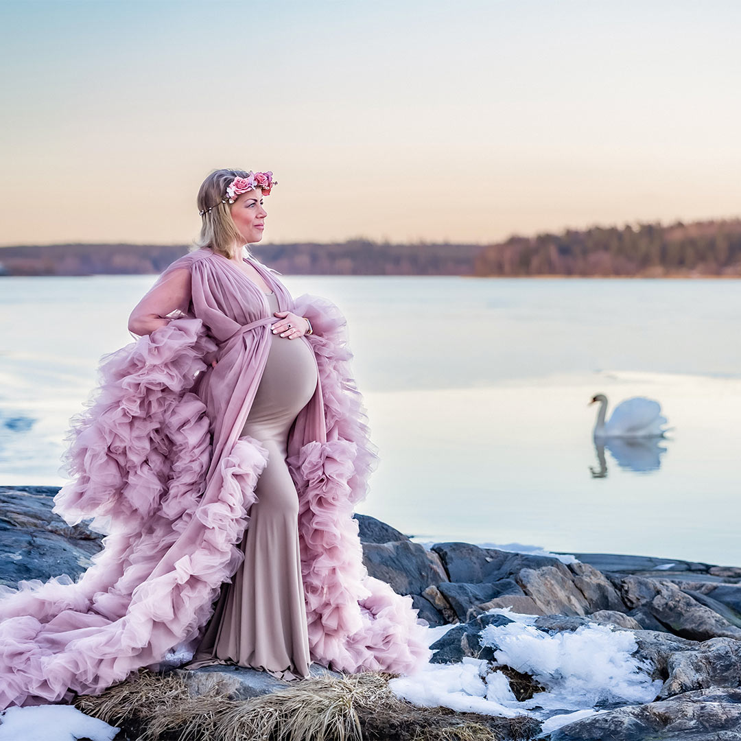 Bilden är från en gravidfotografering en vinterdag med snö på marken, men havet i bakgrunden saknar is och där simmar en svan. I förgrunden står en gravid kvinna med en vacker rosa tyllklänning och rosa blomsterkrans.