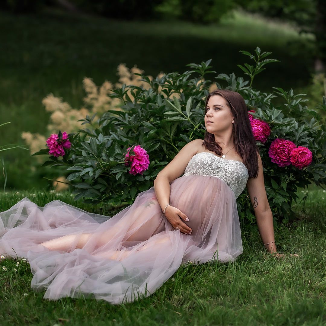 En gravid kvinna i genomskinlig tyllklänning sitter på gräset framför en blommande pionbuske.