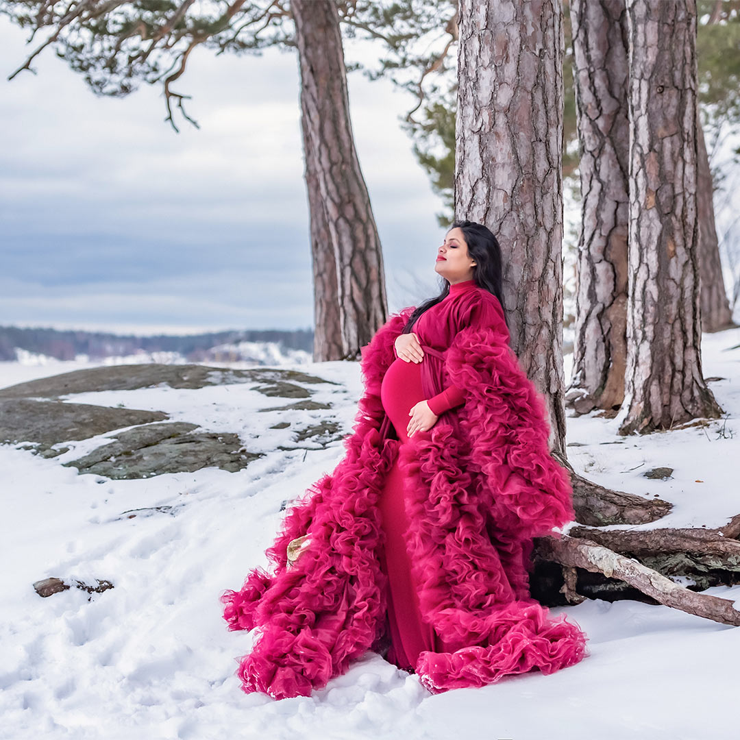 En gravid kvinna klädd i vacker röd tyllklänning lutar sig mot ett träd. Det är en vinterdag med mycket snö.