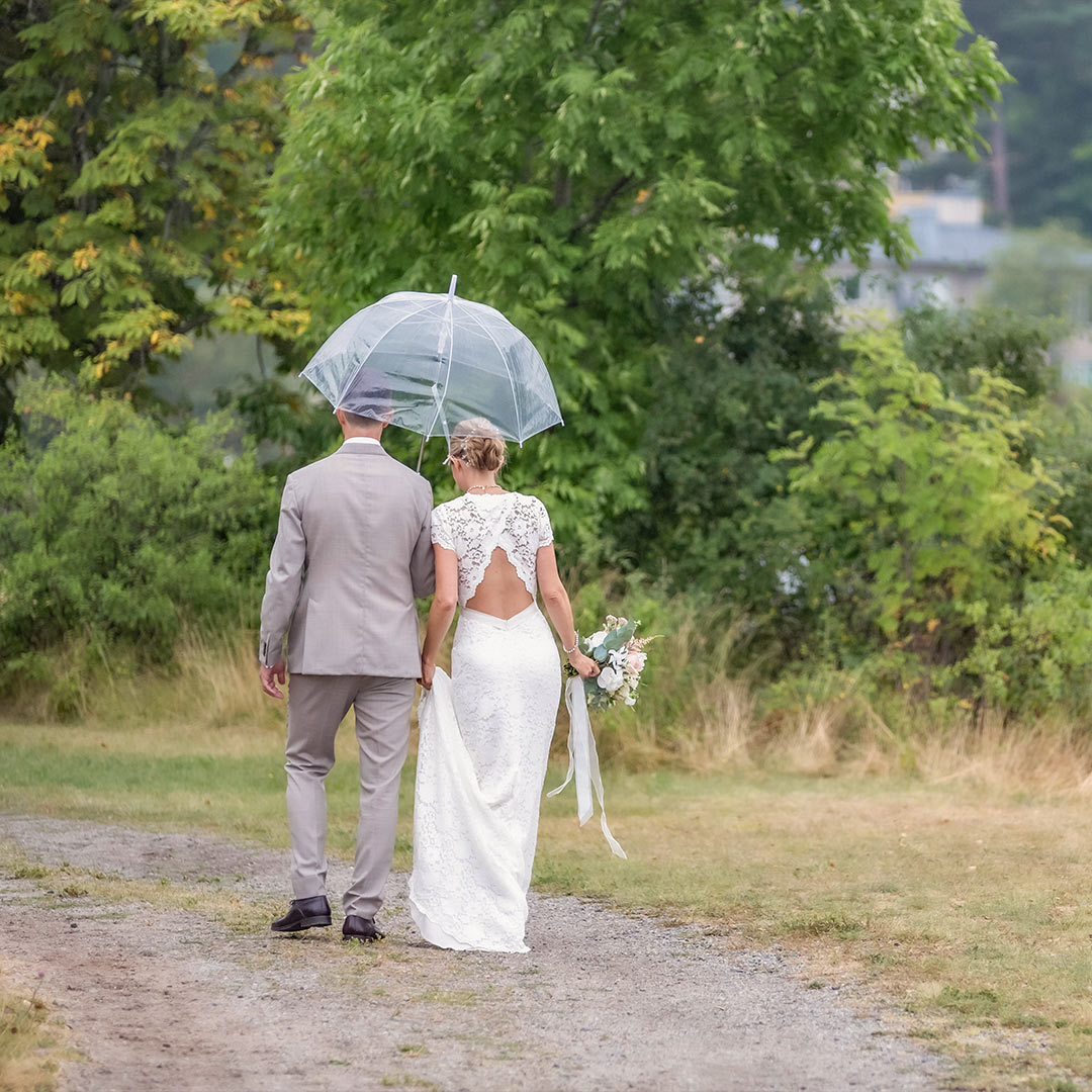 Bröllopspar går på en gång ute i skogen med ryggen mot kameran. Det regnar och de går tillsammans under ett genomskinligt paraply.