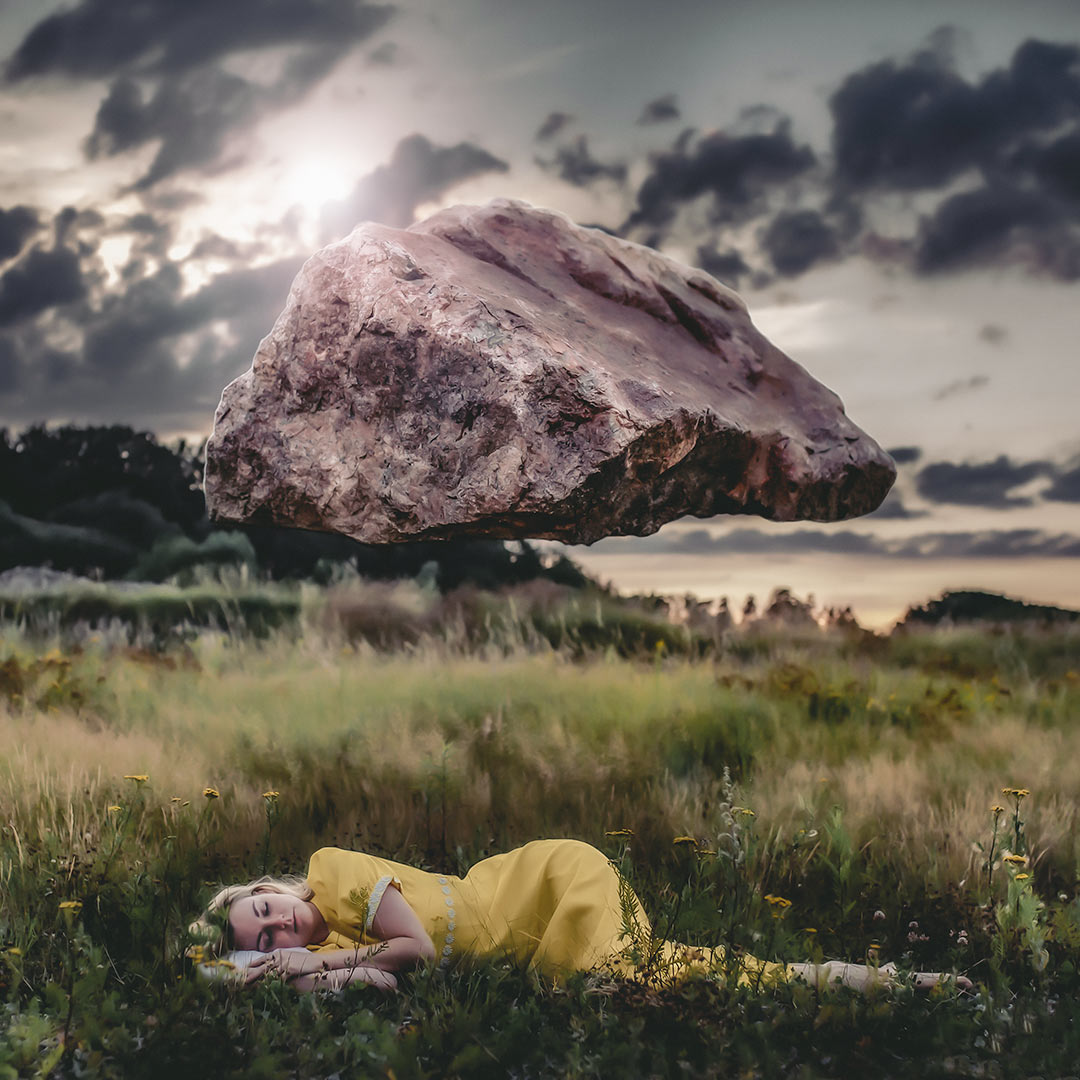 En kvinna i gul klänning sover i gräset. I luften ovanför henne hänger en stor sten.
