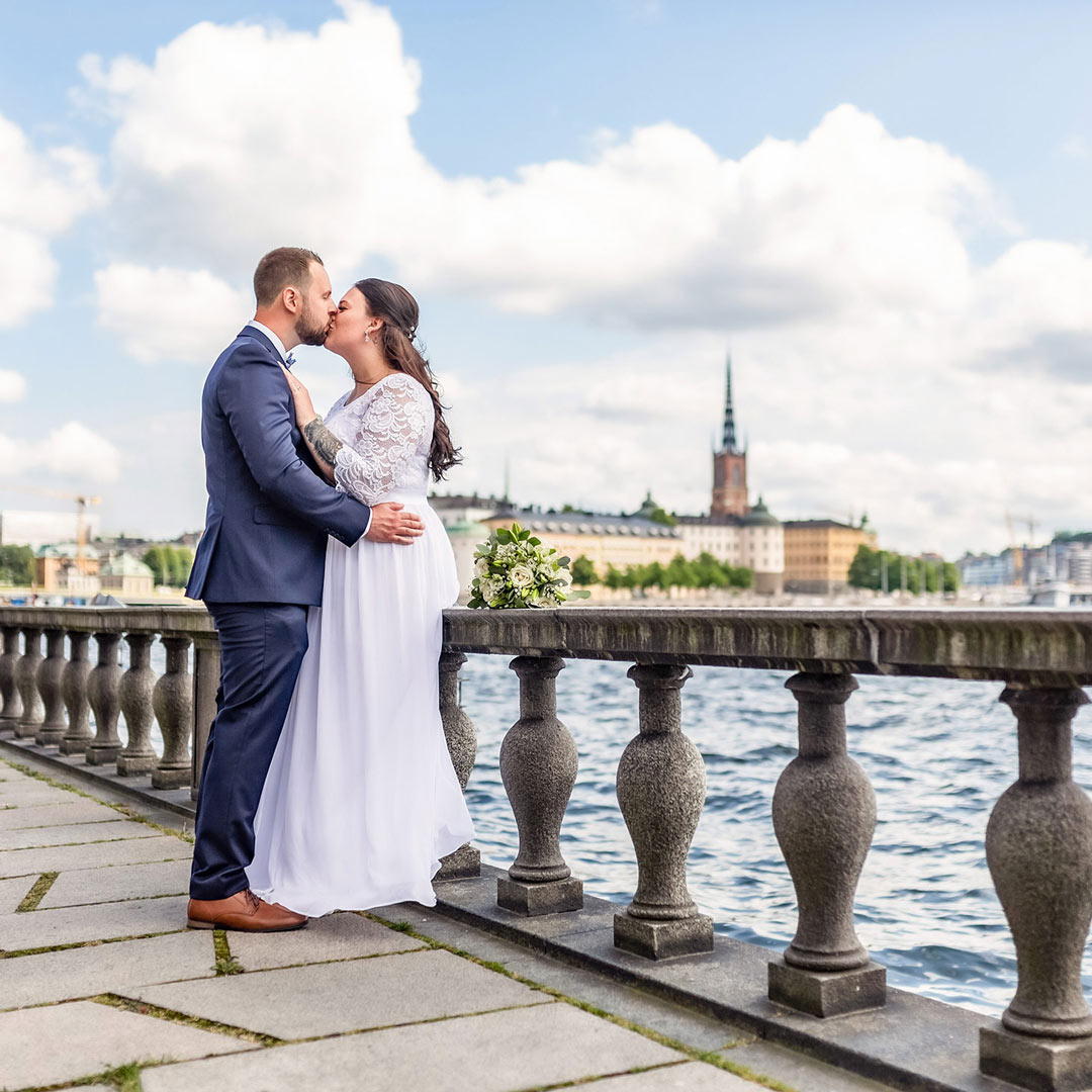 Brudgum och brud pussas vid vattnet. På andra sidan vattnet syns Stockholms Gamla stans siluett.