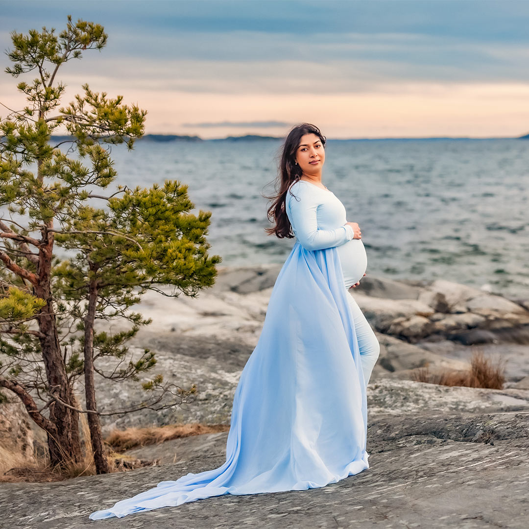 Gravidfotografering av en kvinna i elegant blå klänning med släp. Kvinnan står på klipporna bredvid en liten tall och tittar bakåt mot kameran. I bakgrunden syns havet och solnedgången.