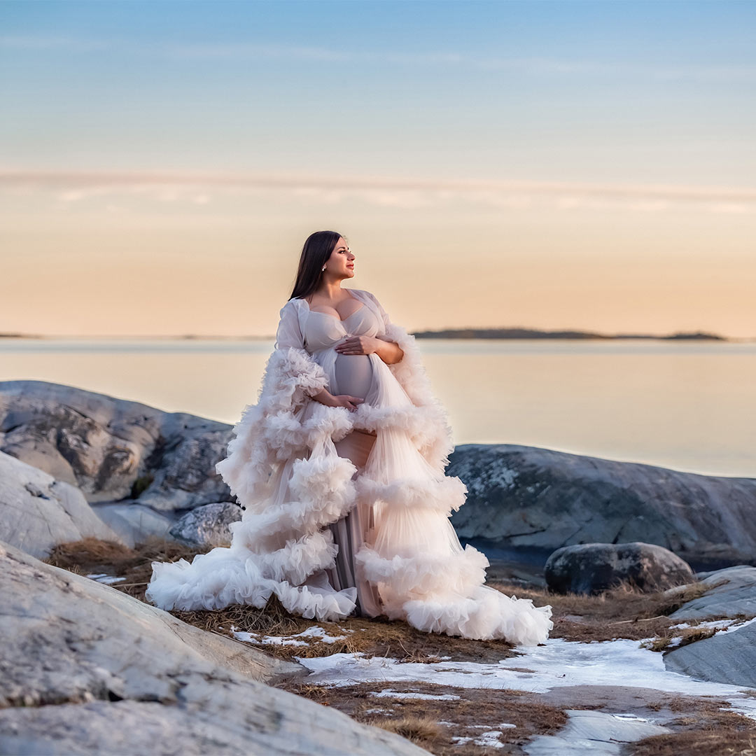 En gravid kvinnan tittar ut över havet och solnedgången. På sig har hon en krämvit tyllklänning. Det ser ut som sommar, men på marken ligger snö.