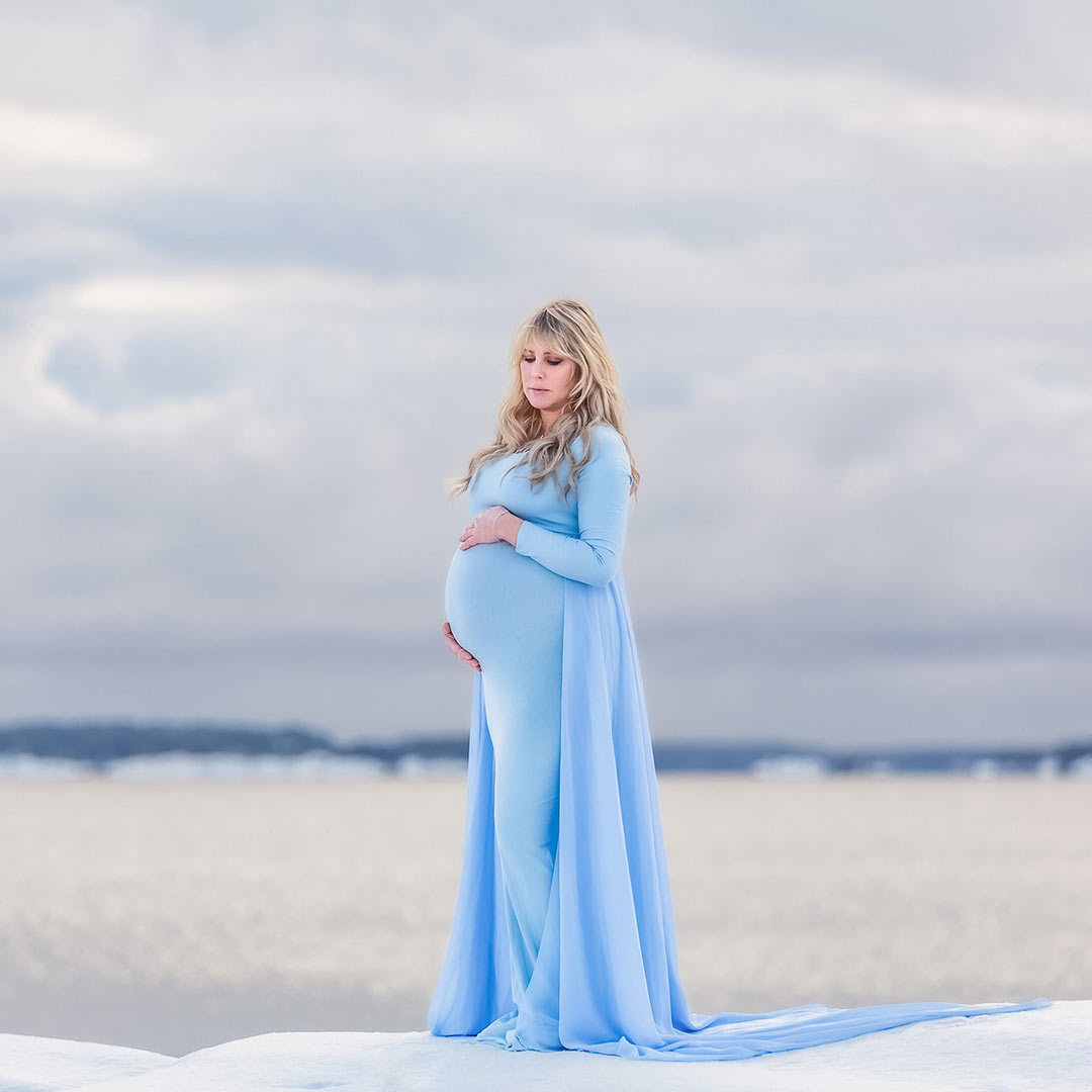 En gravid kvinna i blå gravidklänning med släp står på en höjd som är täckt med snö. Nedanför syns havet som glittrar av solen.