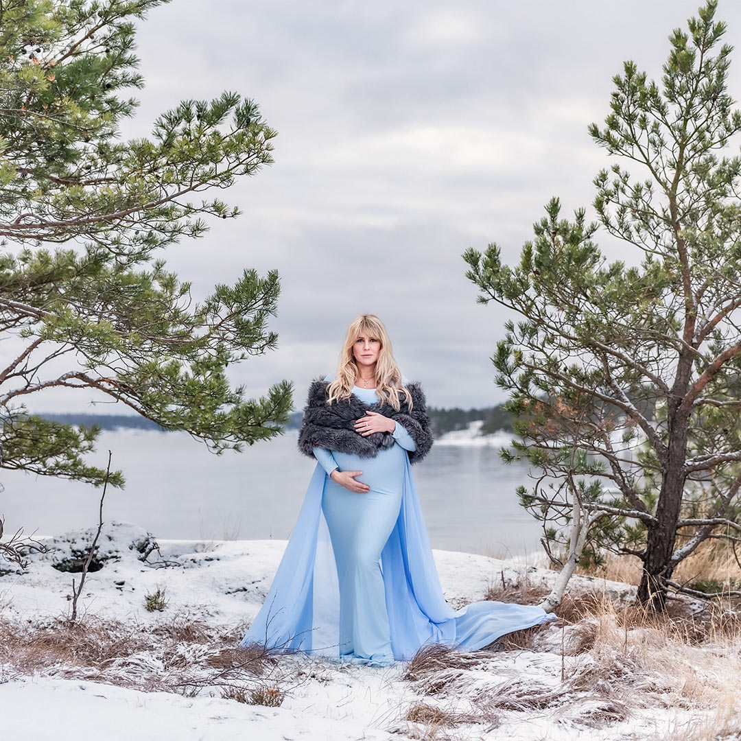 En gravid kvinna i blå klänning står mellan två barrträd i ett snöigt landskap. I bakgrunden syns ett stilla hav.