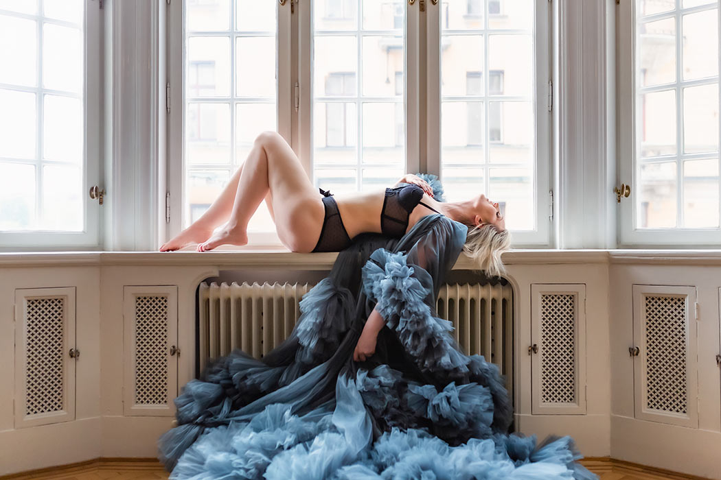 Boudoirfotografering där den vackra kvinnan ligger i ett fönster och har på sig en blå tyllklänning.