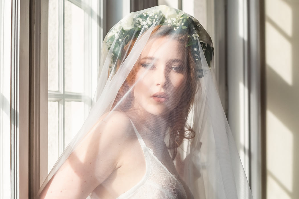 Boudoirfotografering för att skapa en morgongåva. Kvinnan har på sig en blomsterkrans, slöja och vit spets-BH.