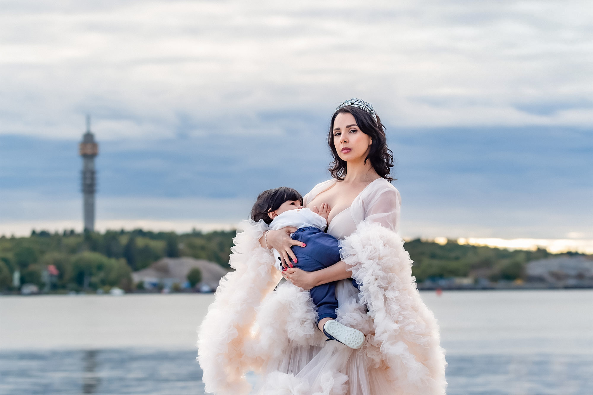 Bild från en Mamma och jag-fotografering. Kvinnan har en vacker krämvit tyllklänning och ammar sitt barn. I bakgrunden skymtar Kaknästornet på andra sidan vattnet.