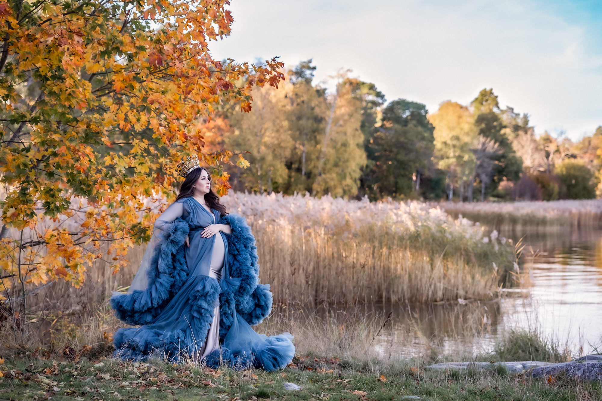 Gravidfotografering om hösten. Den gravida kvinnan har på sig en extravagant blå tyllklänning och en lyxig krona. Hon står på en gräsplätt bredvid ett stort höstträd med gula och orange löv. I bakgrunden syns vatten och ändlöst med vass.