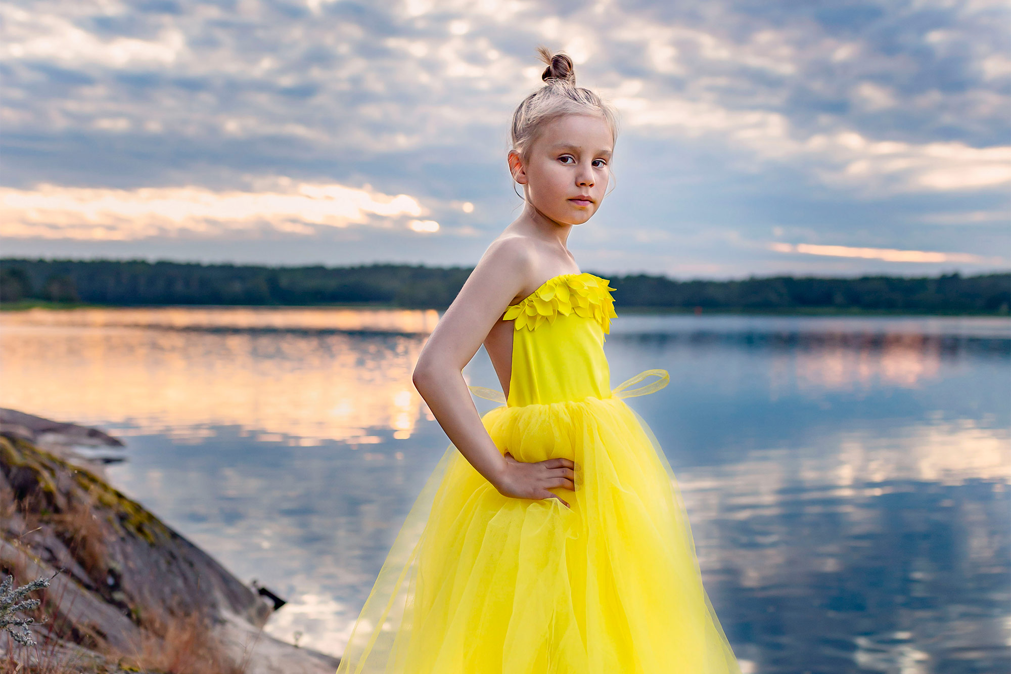 En nioårig flicka i gul tyllklänning står på klipporna vid havet. Flickan tittar in i kameran och i bakgrunden går solen ner och skapar vackra färger i vattnet och himlen.