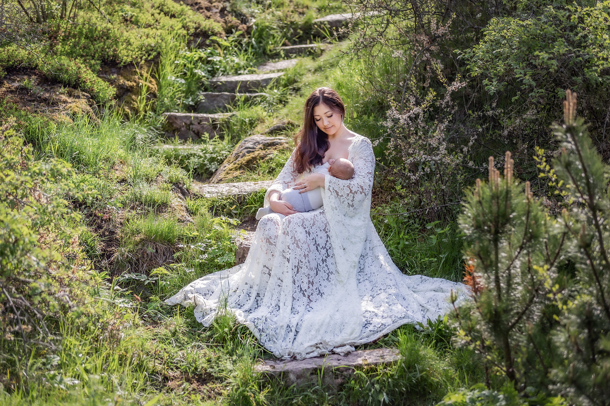 En mamma sitter i en grönskande trappa och ammar sitt barn. På sig har mamman en bohemisk vit spetsklänning och solen skiner ner i trappan.