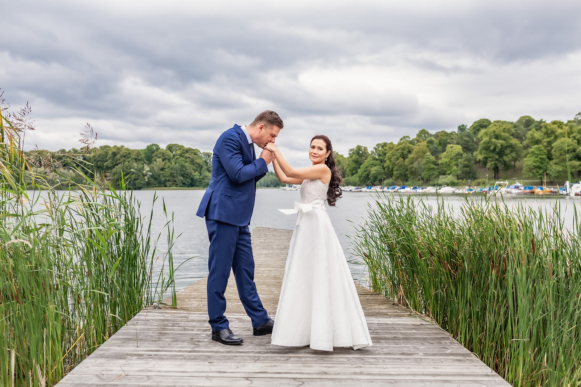 Brudpar står på en brygga vid vattnet vid Stallmästaregården, omgivna av vass. Mannen pussar kvinnan på händerna och kvinnan tittar in i bröllopsfotograf Linda Holmkratz kamera.