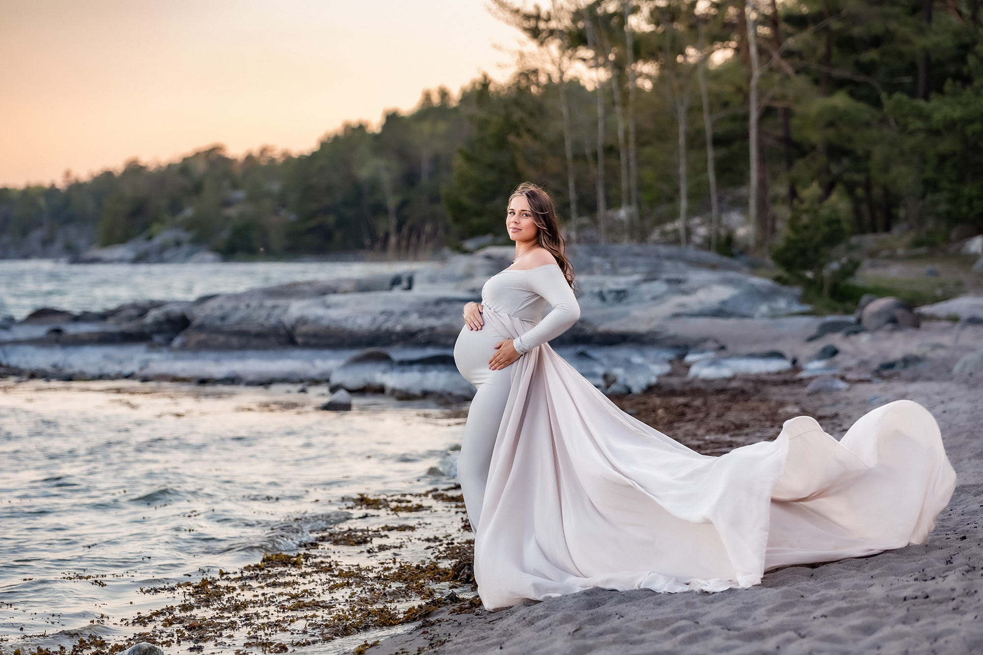 En vacker gravid kvinna står på en sandstrand i en beige gravidklänning som flyger i vinden. I bakgrunden syns klippor och en skog.