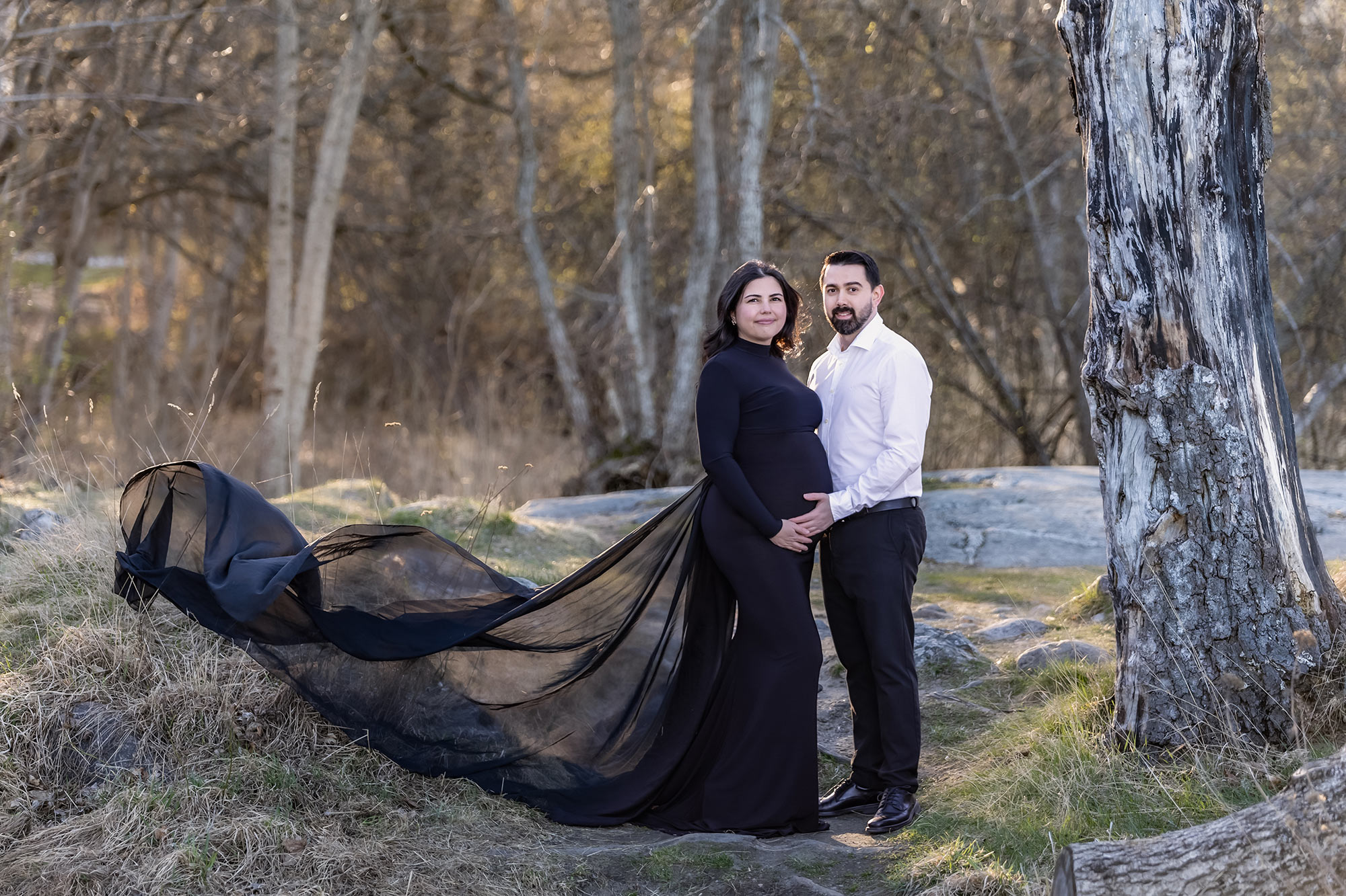 Gravidfotografering med vackert par i stilrent svart och vitt som står på en stig i skogen.