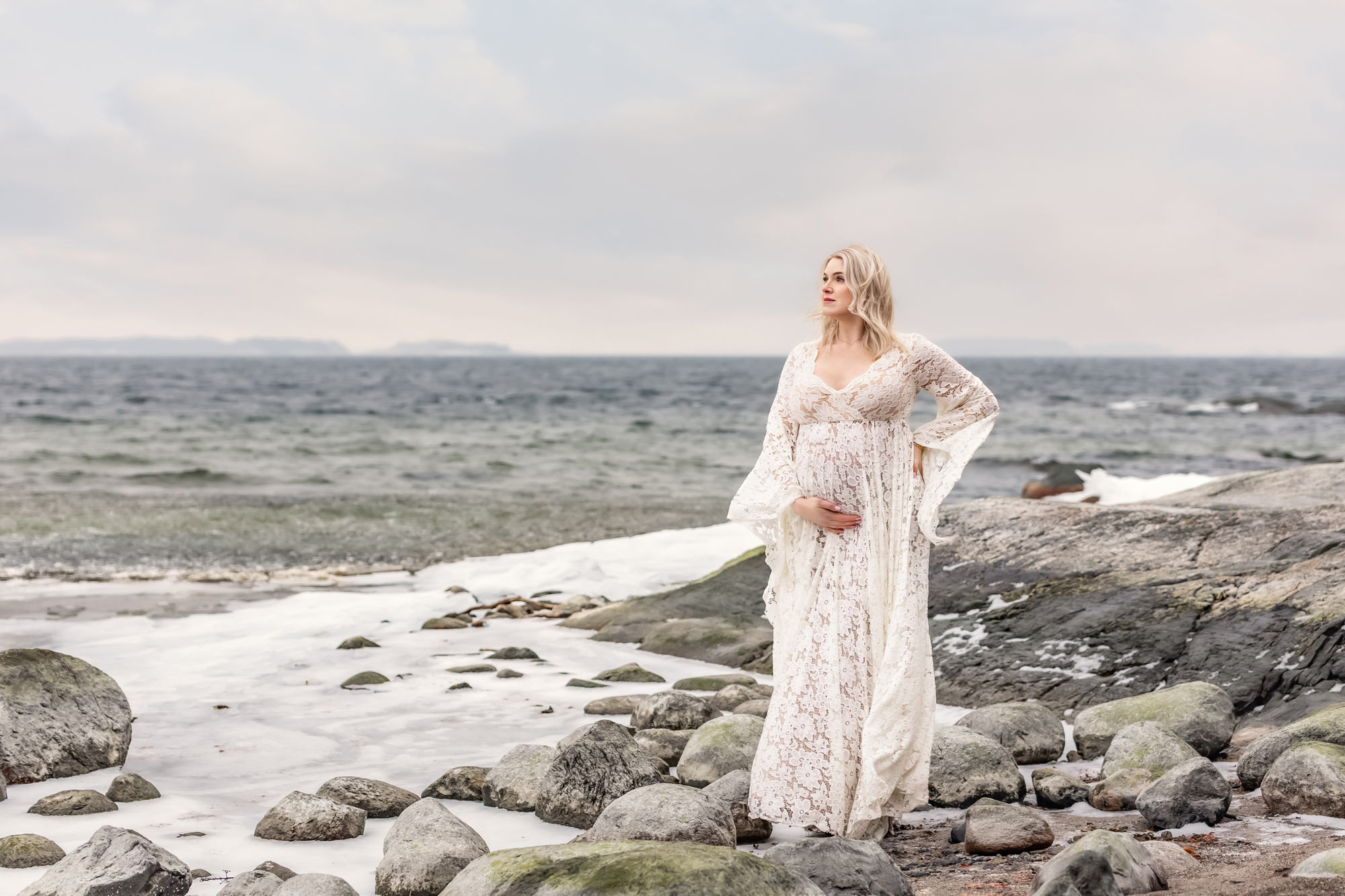 En vacker gravid kvinna i vit spetsklänning står bland klippor och stenar vid havet som delvis är täckt av is.