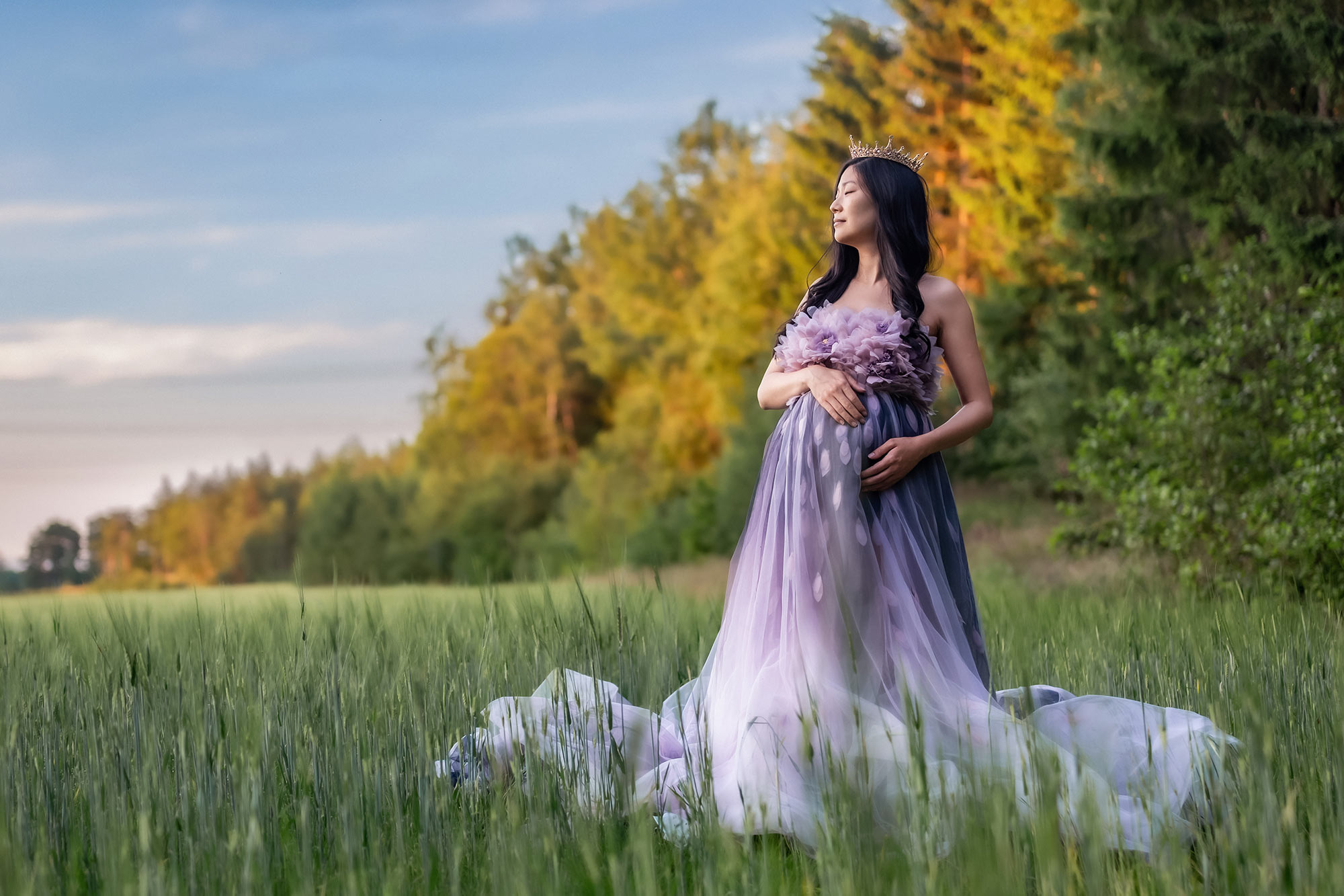 Gravidfotografering i värme en högsommardag. Den gravida kvinnan står vid en äng och har på sig en fantastisk gravidklänning med långt släp som ser ut som en blomma.