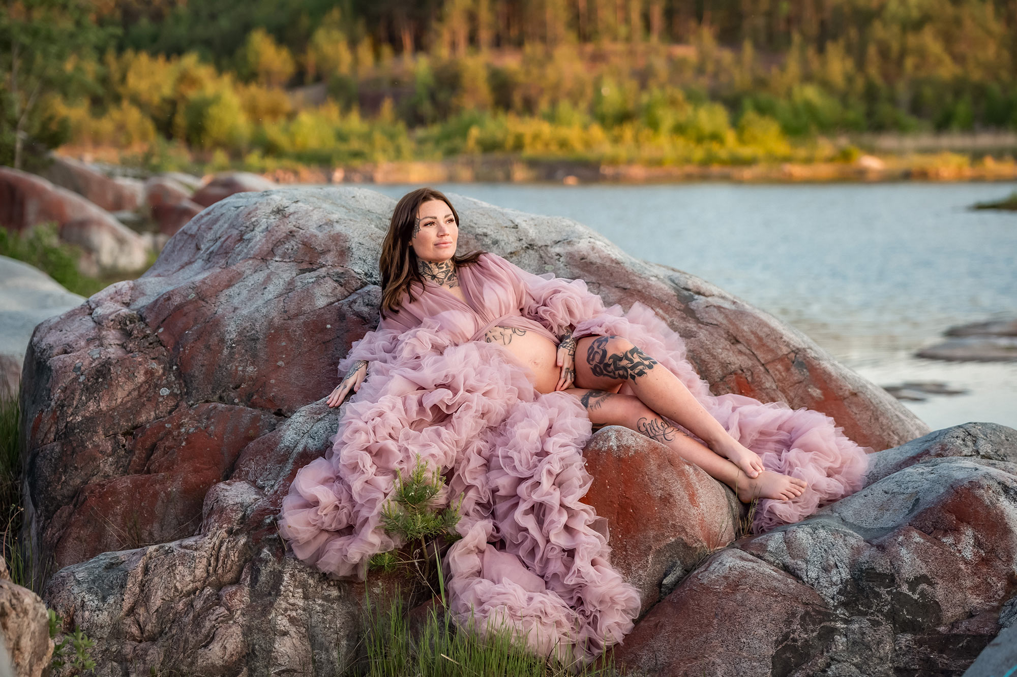 Gravidfotografering bland klipporna i ett stenbrott. I bakgrunden syns vatten och en skog i kvällsljus. Kvinnan har på sig en rosa gravidklänning i tyll som flöder ut över klipporna.