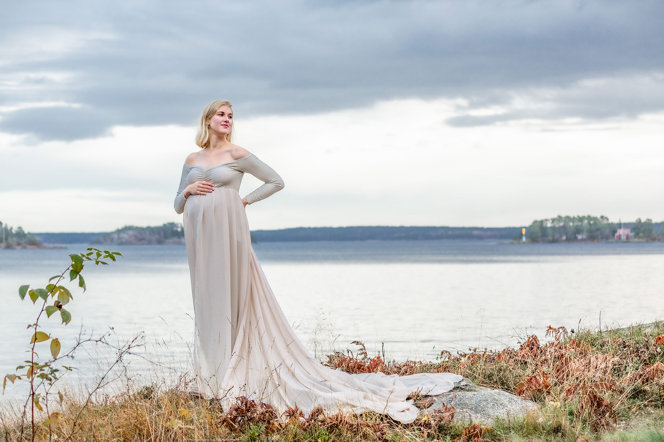 Gravidfotografering i kyla. Det är november och den gravida kvinnan har en beige gravidklänning med långt släp. Hon står på en höjd och i bakgrunden syns havet sträcka ut sig.