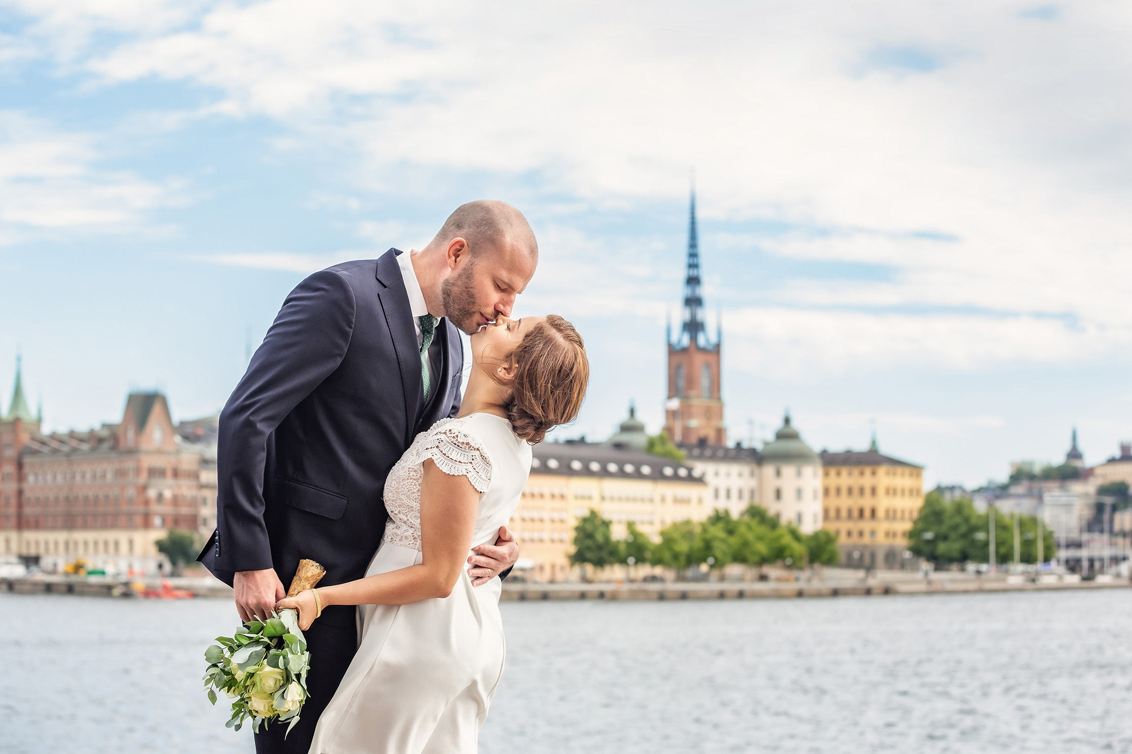 Nygift par blir fotograferat när de pussas vid vattnet i närheten av Stockholms stadshus. I bakgrunden syns gamla stans vackra gamla byggnader.