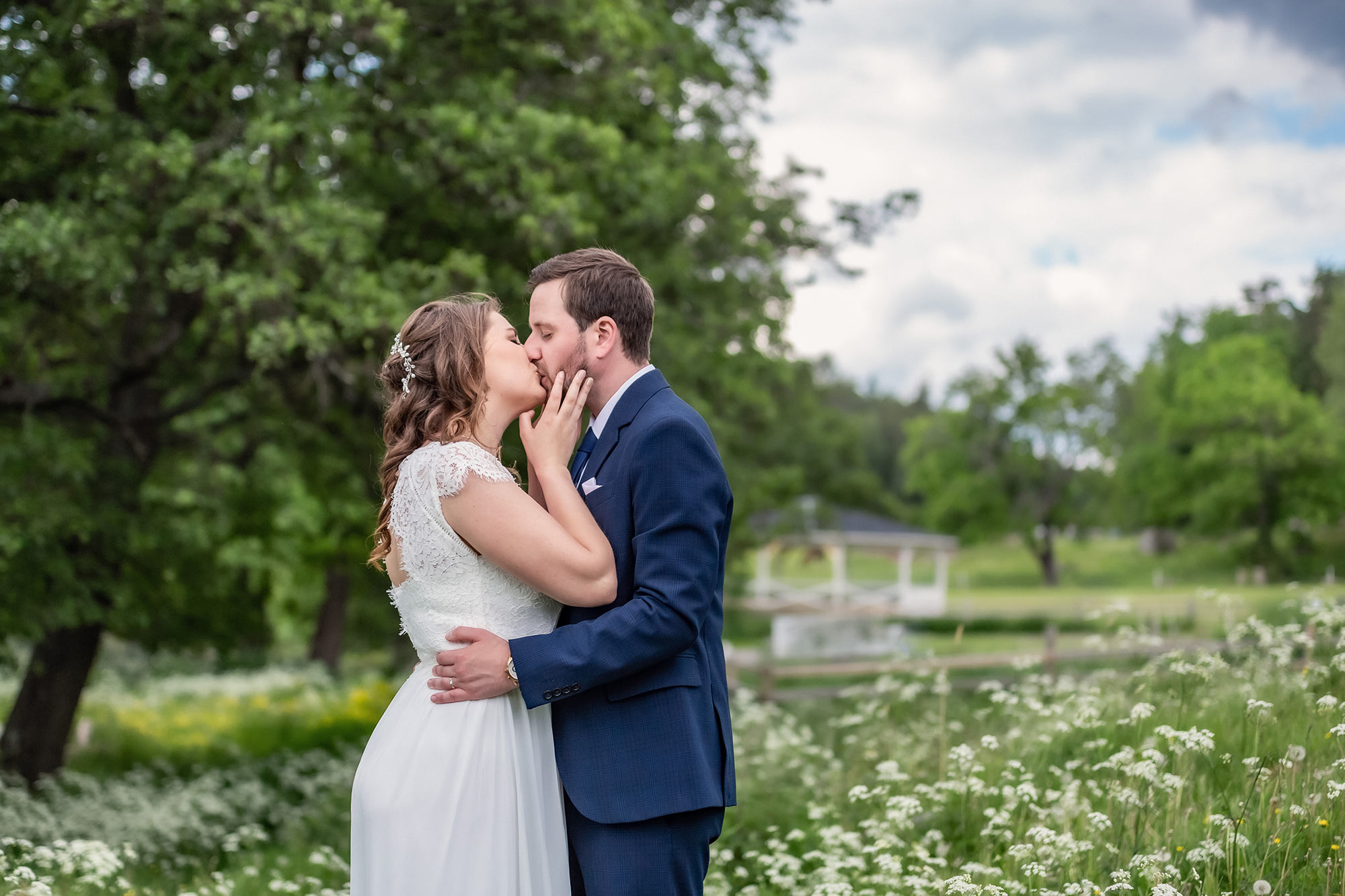 Bröllopspar pussas på en vacker sommaräng. I bakgrunden syns träd och en dansbana.