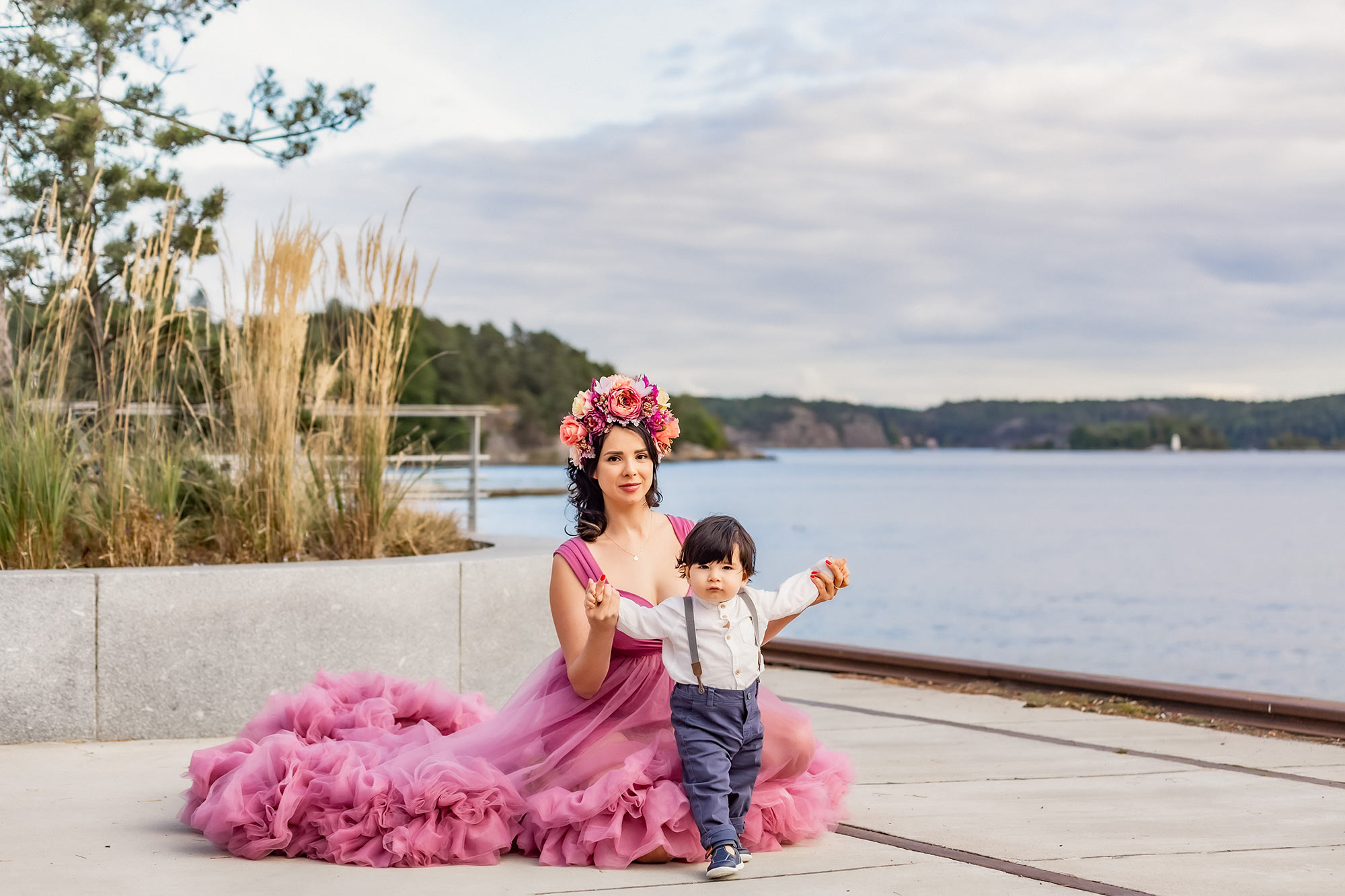 En mamma i rosa tyllklänning och med en blomstertiara sitter på en kaj och håller sin lilla son i händerna för att hjälpa honom att ta sina första steg. I bakgrunden syns havet och grönska.