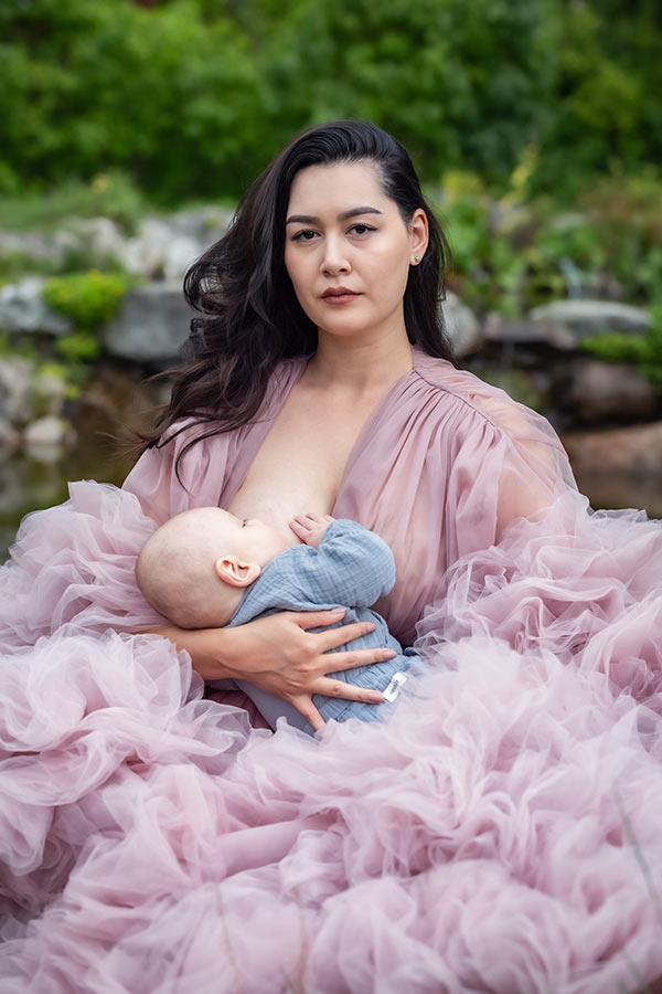 En mamma sitter i naturen och ammar sitt barn. Hon har en rosa tyllklänning på sig och tittar in i kameran.