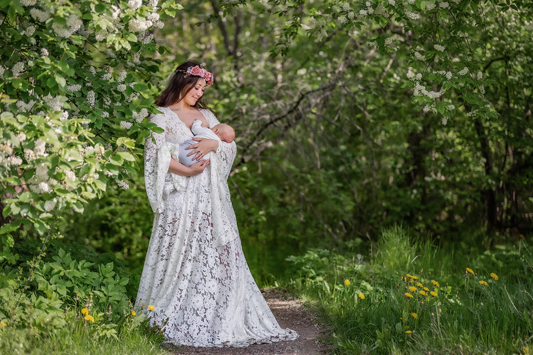 Amningsfotografering där kvinnan har en vit spetsklänning och står vid vitblommande grönskande träd och ammar sin bebis.