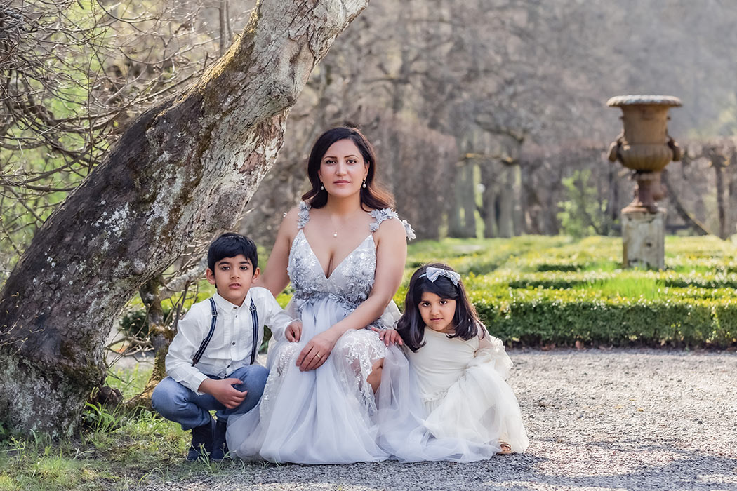 Familjefotografering med en mor och hennes två barn. De sitter på huk i en slottsträdgård.