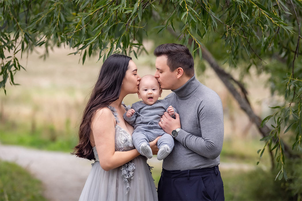 Familjefotografering i september under ett pilträd. Familjen har grå kläder och mamman och pappan håller barnet mellan sig och pussar barnets panna.