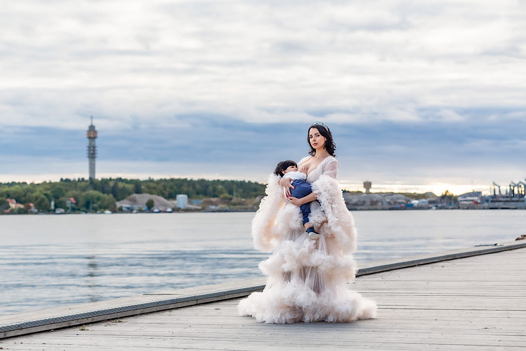 Bild från en Mamma och jag-fotografering. Kvinnan har en vacker krämvit tyllklänning och ammar sitt barn. I bakgrunden skymtar Kaknästornet på andra sidan vattnet.