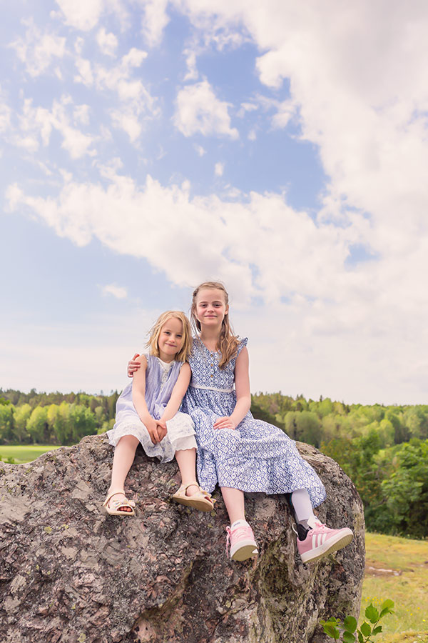 Två systrar i blå sommarklänning sitter på en stor sten uppe på en höjd. I bakgrunden syns en grön skog lite uppifrån och på himlen flyter vita moln fram.