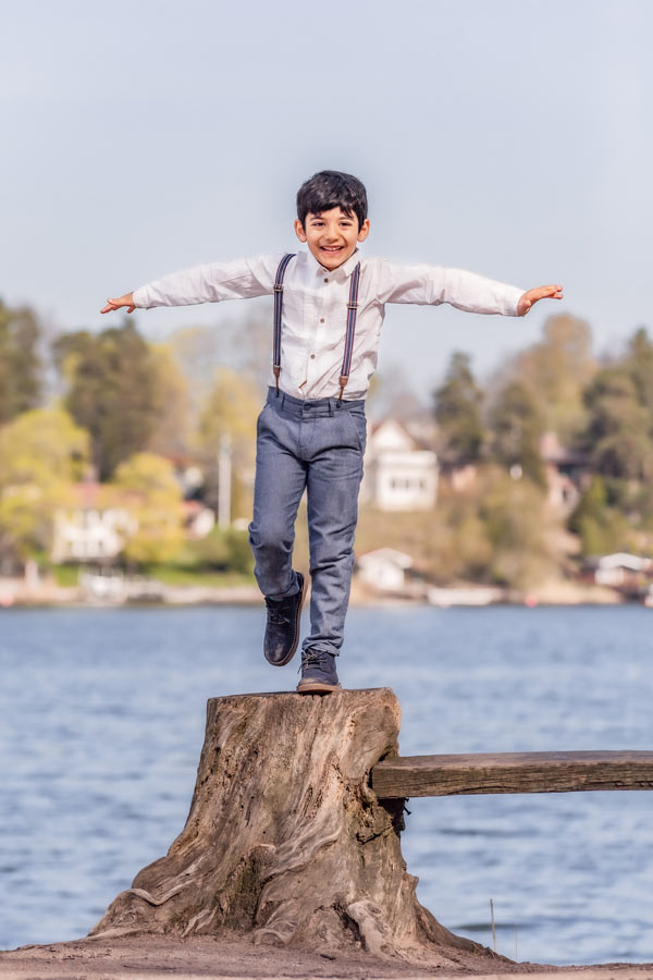 En pojke står på ett ben på en stor stubbe vid vattnet.
