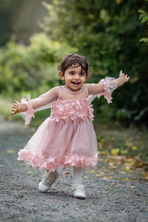 Ett litet barn i rosa klänning springer på en grusgång med armarna utslagna och skrattar in i kameran.