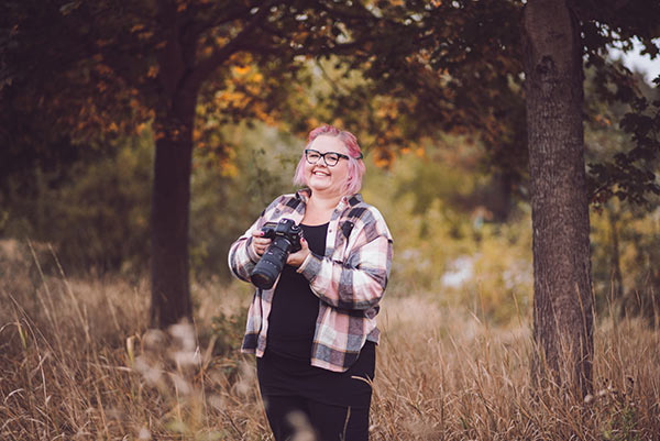 Fotograf Linda Holmkratz står på en äng med höga gräs och träd. Hon skrattar och håller i sin kamera.