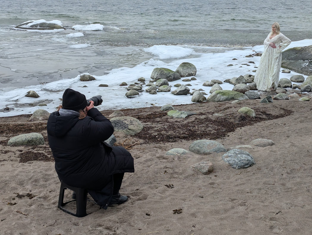 Gravidfotograf Linda Holmkratz fotograferar en gravid kvinna på stranden bland stora stenar vid havet. Bilden är tagen av den blivande pappan.