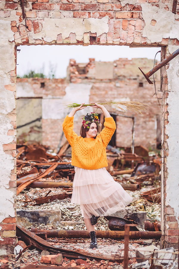 En kvinna med tyllkjol och orange tröja står i en dörröppning i en ruin av ett tegelhus. Himmelen skymtar i bakgrunden och på marken ligger rostigt skrot.