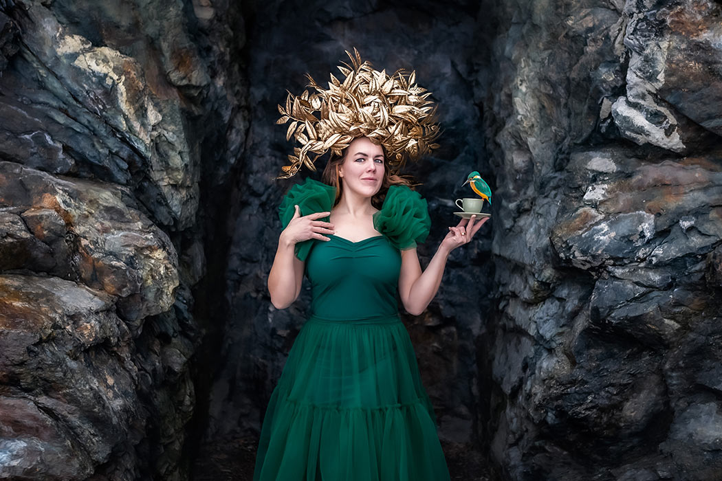 En kvinna i grön tyllklänning och med en stor guldkrona på huvudet, står mellan två klippor och håller en grön kaffekopp. På koppen sitter en lite grön och orange fågel.